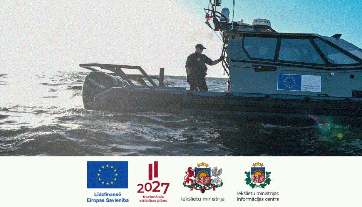 Fotoattēls, kurā redzams Latvijas robežsargs uz jūrā esoša Valsts robežsardzes kutera klāja, kā arī Eiropas Savienības līdzfinansējuma, Nacionālā attīstības plāna, Latvijas Republikas Iekšlietu ministrijas un Iekšlietu ministrijas Informācijas centra logotipi