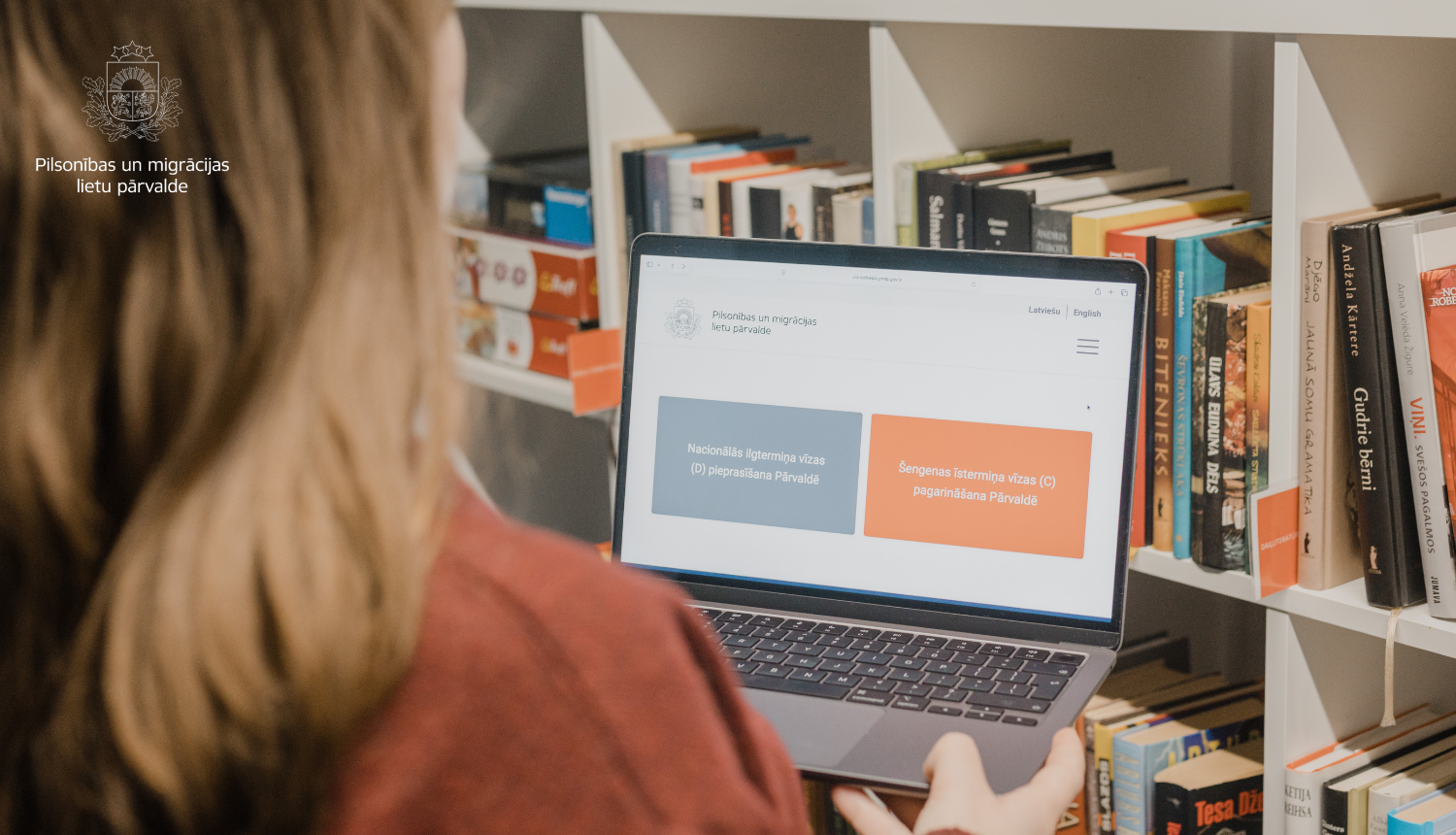 Persona pie grāmatu plaukta skatās portatīvajā datorā, kur atvērta Pilsonības un migrācijas lietu pārvaldes mājaslapa ar vīzas pieteikšanas pogām.